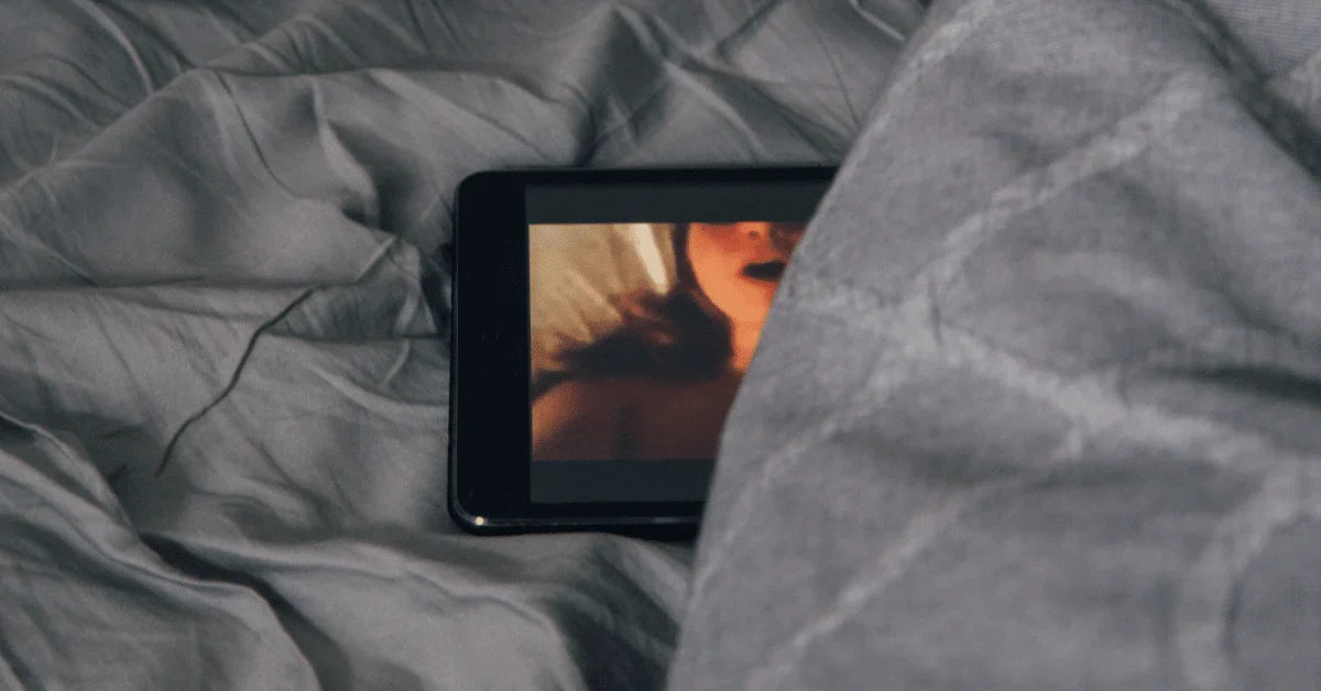 Tablet on bed partially covered by grey blanket, displaying intimate video content