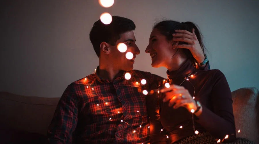Romantic couple smiling on a couch, wrapped in warm string lights indoors