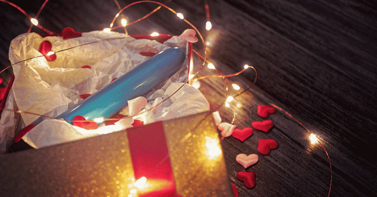 Gift box with a blue adult toy, heart confetti, and festive string lights on wood surface