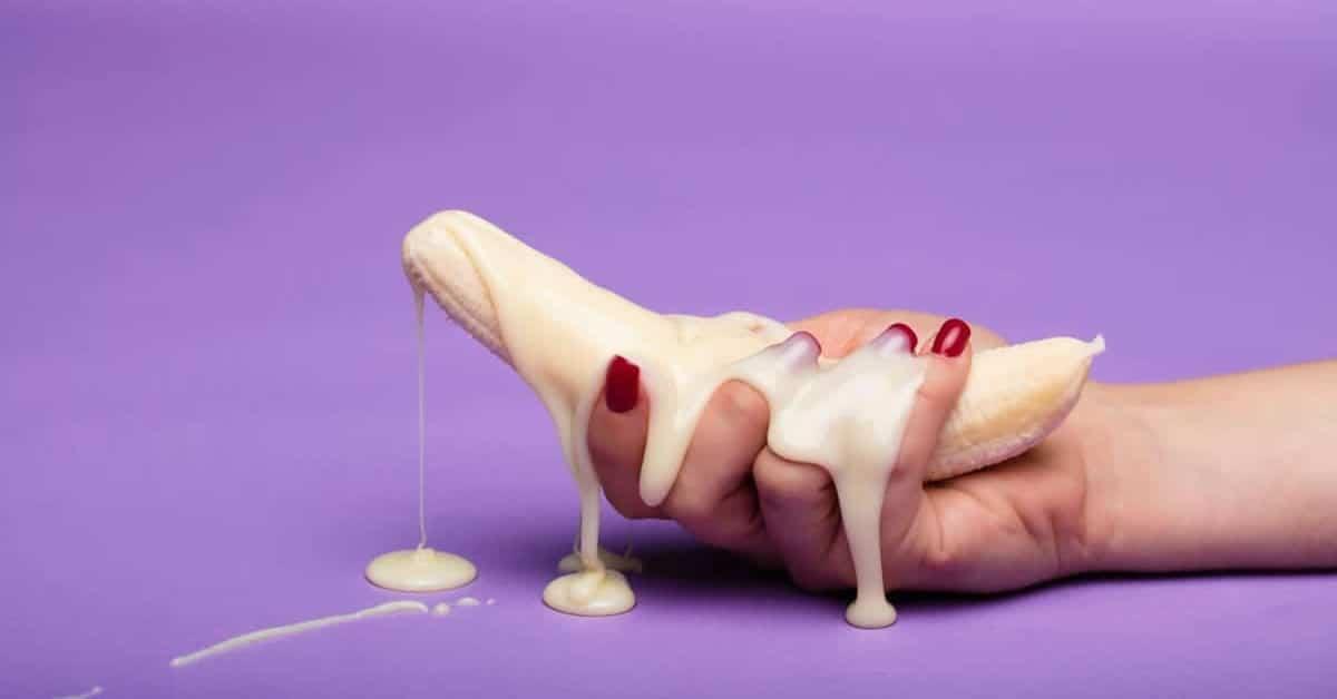 Hand holding a peeled banana with creamy sauce dripping, purple background, sensual food concept