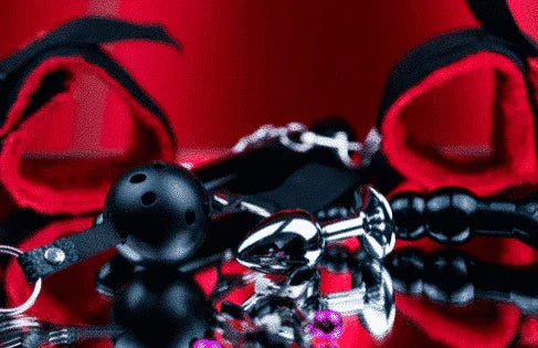 Svart ballgag, metallisk analplug og røde bånd på rød overflate – erotisk leketøy for par