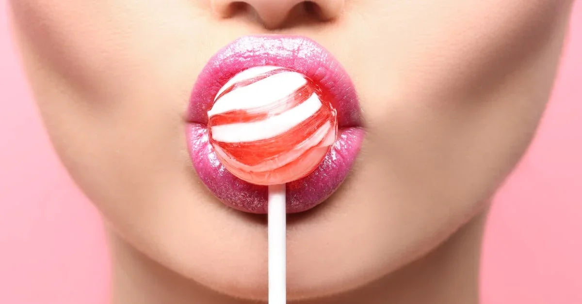 Close-up of glossy pink lips holding a red and white lollipop against a pink background.
