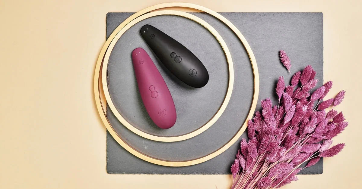 Two Womanizer stimulators in black and burgundy on slate with pink dried flowers