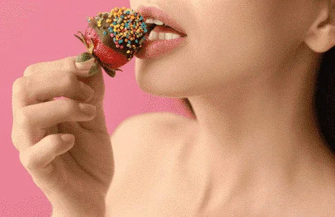 Kvinne spiser sjokoladejordbær med fargerike strøsler mot rosa bakgrunn
