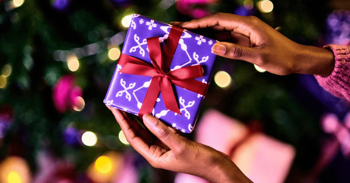 Hands holding a wrapped Christmas gift with red ribbon in front of a festive tree