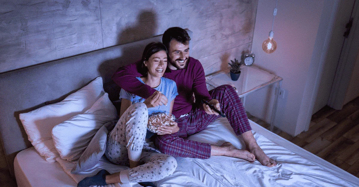 Happy couple in pajamas relaxing in bed, watching TV and sharing popcorn in a cozy bedroom