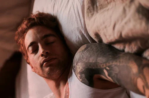 Tattooed man with brown hair sleeping on bed, relaxed in soft bedroom lighting.