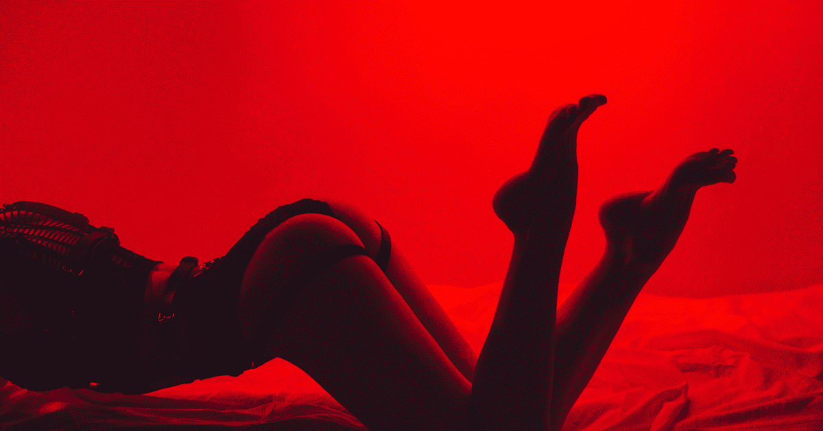 Person i undertøy som ligger på en seng med rødt lys, sensuell stemning og intim atmosfære.