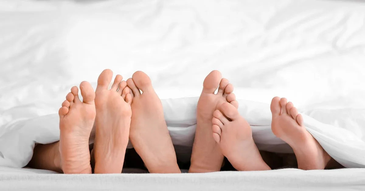 Tre par føtter under en hvit dyne, som antyder intimitet i sengen.