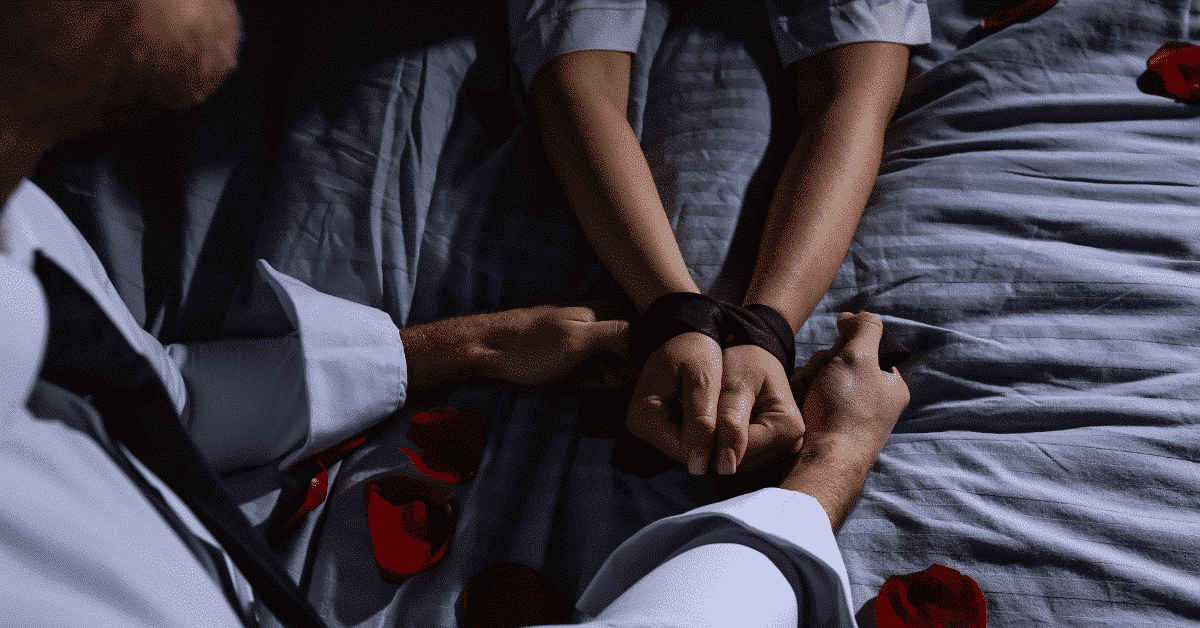 Couple on bed with hands tied using soft restraints, rose petals scattered, intimate setting