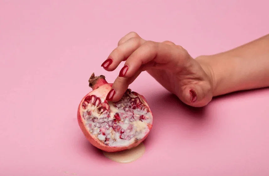Hånd med rød neglelakk berører åpen granatappel på rosa bakgrunn