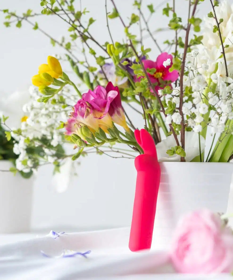 Pink silicone rabbit vibrator displayed beside fresh spring flowers in white pot, bright background