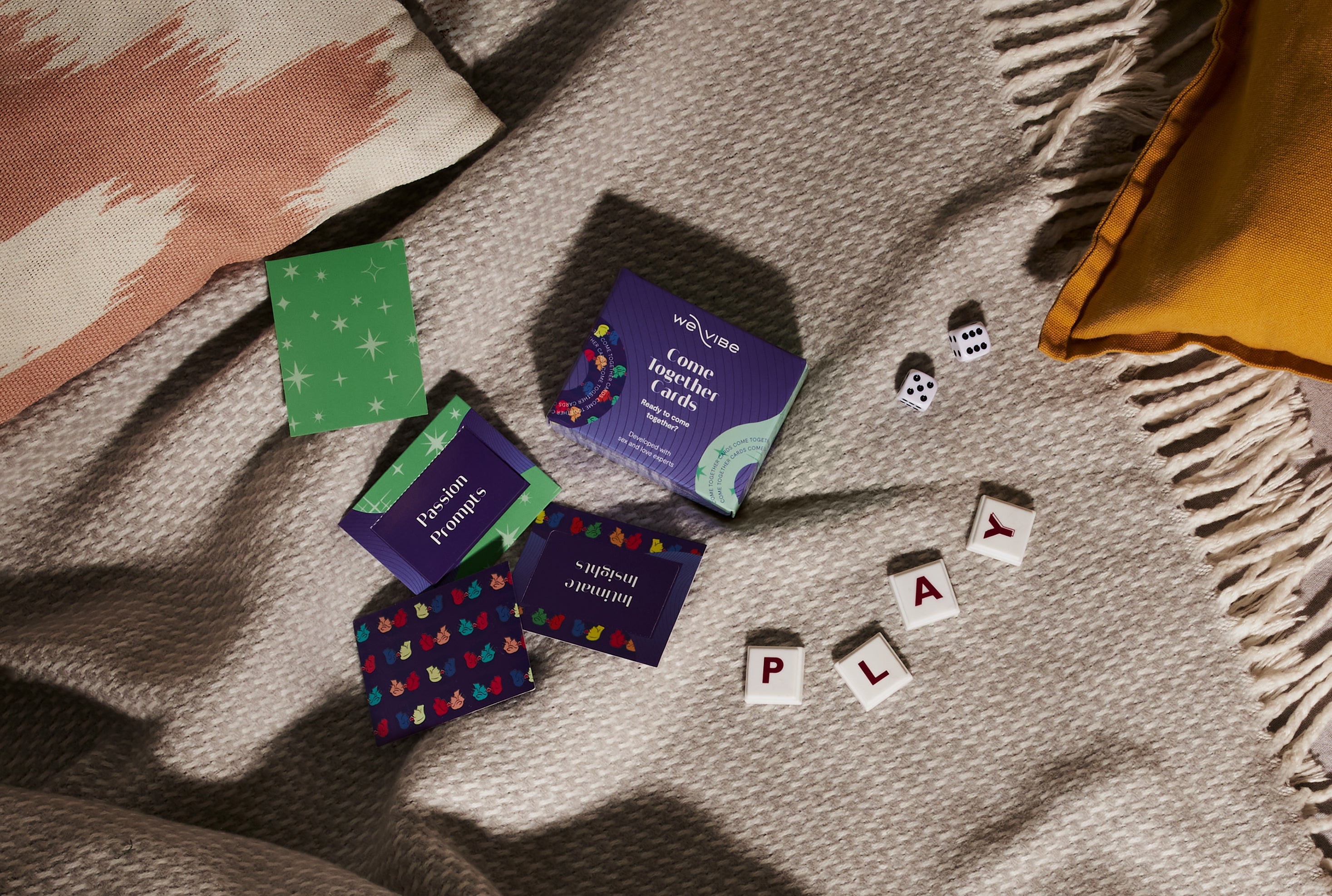 Intimacy card game set, dice, and letter tiles spelling PLAY on cozy blanket with pillows
