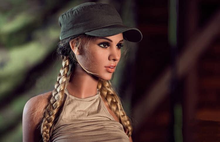 Realistic female doll with long blonde braids and a green cap, wearing a beige top