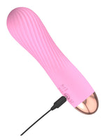 Cuties rosa minivibrator liggende diagonalt med USB-ladekabel koblet til ladeporten i bunnen mot hvit bakgrunn.
