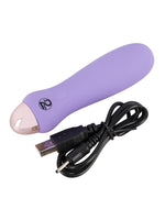 Cuties minivibrator i lilla med medfølgende svart USB-ladekabel liggende ved siden av hverandre mot hvit bakgrunn.