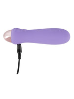 Cuties minivibrator i lilla med USB-ladekabel koblet til ladeporten i bunnen, mot hvit bakgrunn.