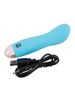 Cuties blå minivibrator med medfølgende svart USB-ladekabel liggende ved siden av hverandre mot hvit bakgrunn.