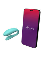 We-Vibe Sync Lite parvibrator i turkis silikon ved siden av smarttelefon som viser We-Vibe-appen, som illustrerer appstyring og fjernkontroll av vibrasjoner.